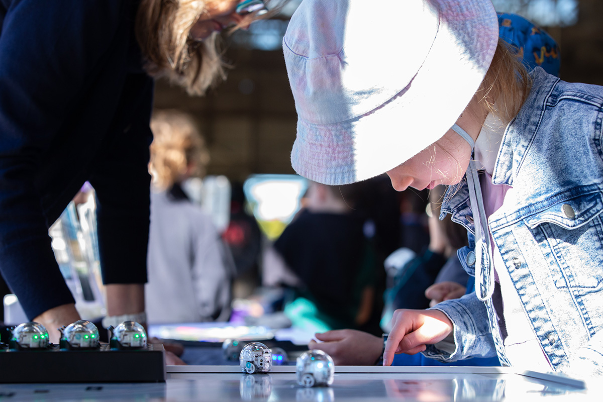 A young child wearing a white hat and denim jacket is focused on interacting with a small robot on a table. An adult stands nearby, assisting other children in the background. The scene appears to be an educational or interactive event involving robotics